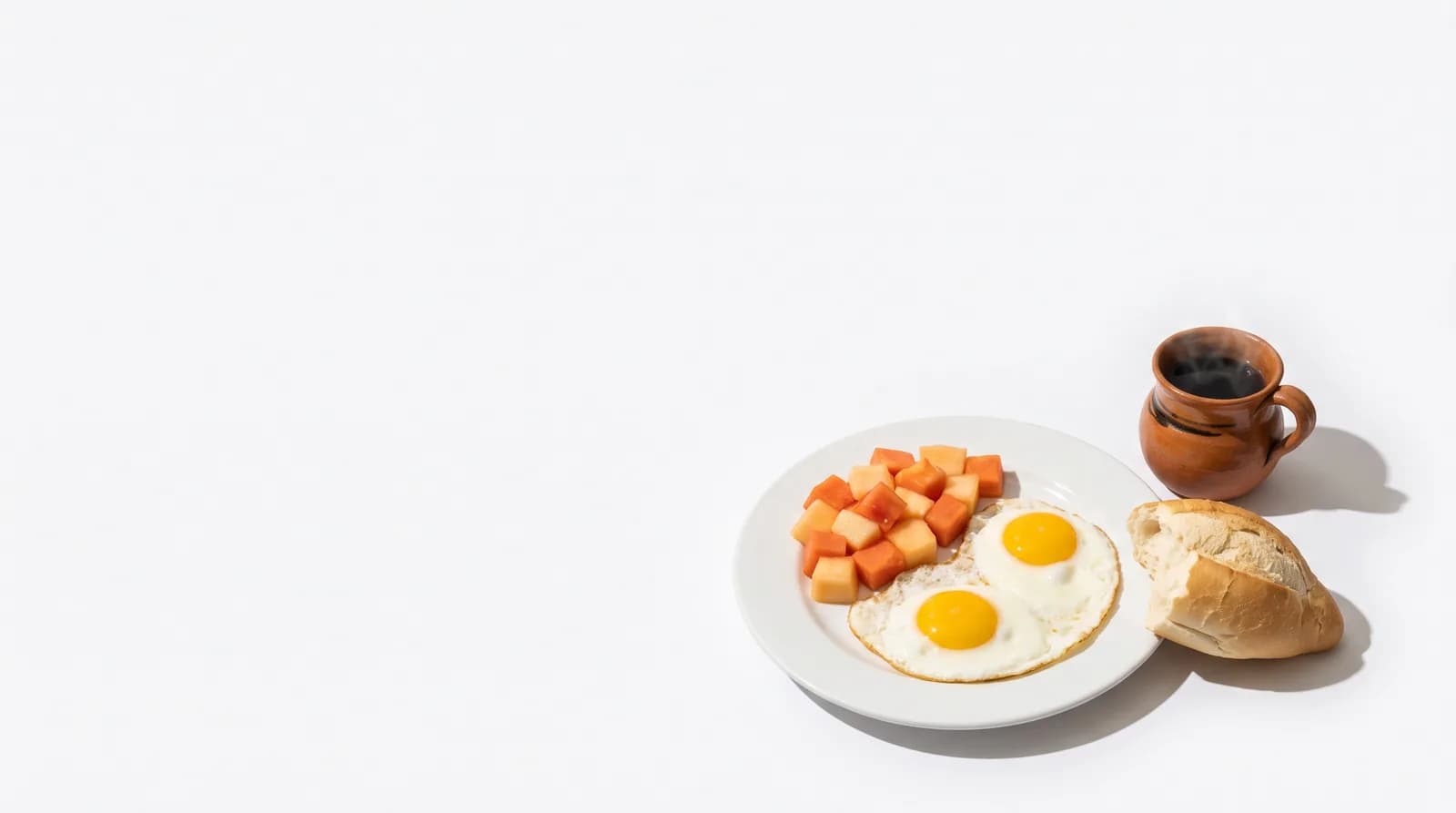 Un plato con huevos estrellados, fruta picada, un bolillo partido y una taza de café.